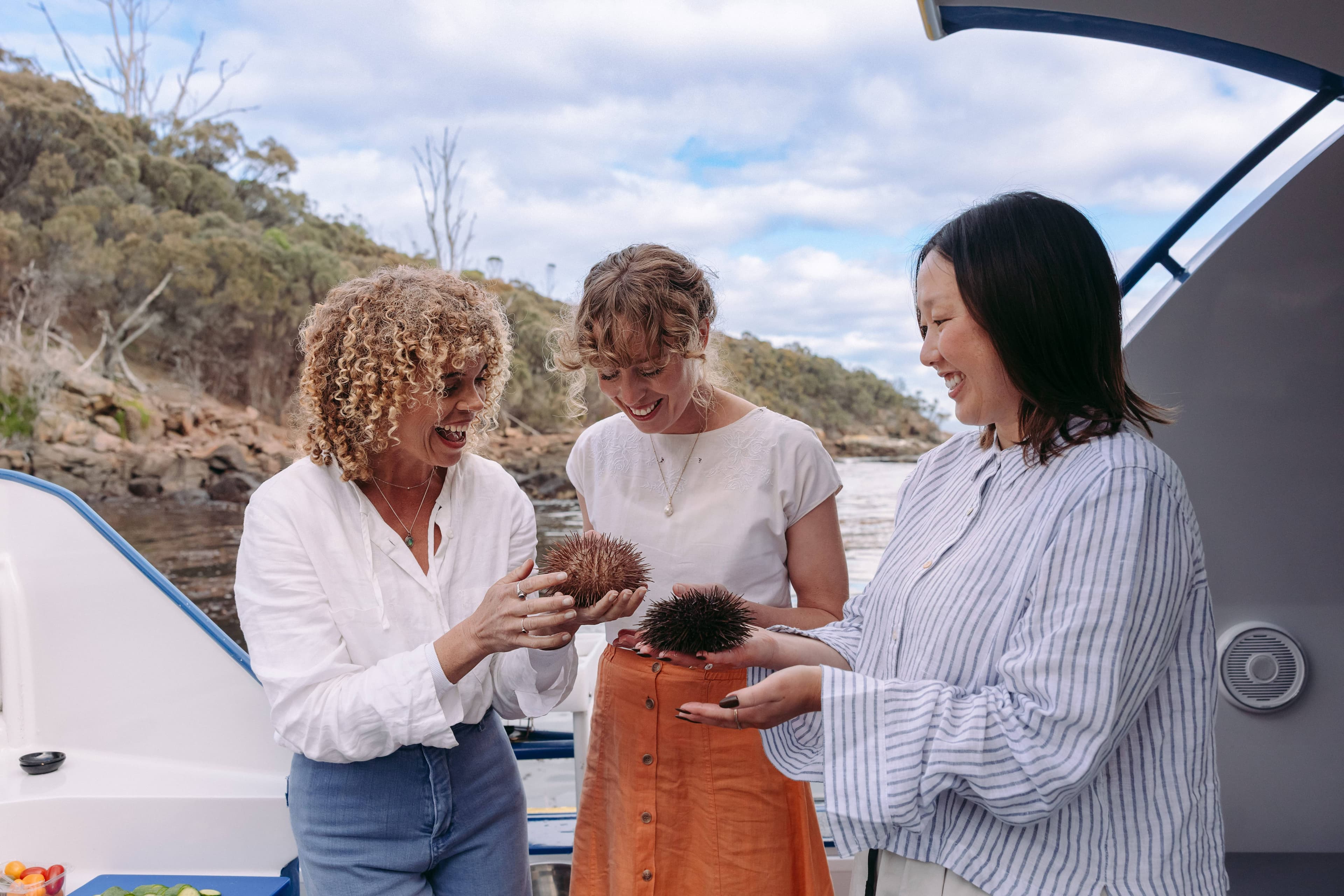 Three women laughing together on a boat, holding sea urchins during a coastal adventure in Australia